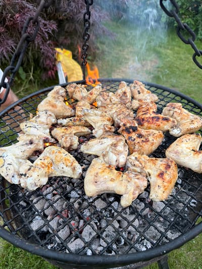 In the city of Mordarka, Poland, a black metal grill hovers over glowing charcoal, with flames licking upwards. On the grill, a generous assortment of chicken wings is being cooked. Some of the wings are pale and lightly browned, while others are well-charred and crispy, indicating they are nearing completion. The grill is suspended by thick black chains, suggesting an outdoor cooking setup, possibly in a backyard or park. In the background, blurred foliage and a yellow bag are visible, hinting at an outdoor gathering or event. The scene is captured at an eye-level perspective, focusing on the food and the cooking process.