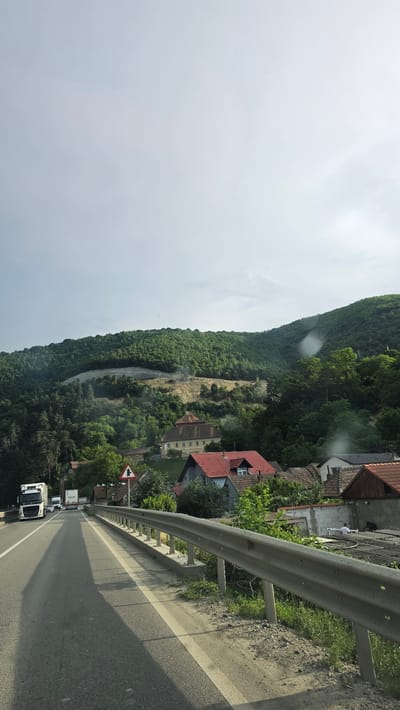 The image shows a road winding through a mountainous, forested area, likely in Boița, Romania. A white truck is driving on the asphalt road, which has a white line marking the lanes. A metal guardrail runs along the side of the road, separating it from a steep embankment covered in lush green trees. On the embankment, there are several houses with red tile roofs, nestled among the foliage. A dirt slope is visible on the mountainside, possibly indicating a recent landslide or construction. A triangular road sign with a black arrow pointing right is also present. The sky is overcast, suggesting a cloudy or hazy day. The overall scene depicts a rural, mountainous landscape with a road cutting through it, and hints of human habitation integrated into the natural environment.