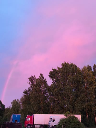 The image depicts a serene evening scene with a vibrant sky painted in hues of blue, pink, and purple. A rainbow arcs gracefully across the left side of the sky. In the foreground, a line of lush green trees stands tall. Below the trees, a red truck and a white refrigerated truck are parked side-by-side. To the left of the trucks, a blue container is visible. The overall atmosphere is peaceful, suggesting a tranquil moment in Brodi, Latvia, during dusk.
