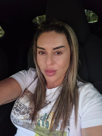 A woman with long blonde and brown hair is sitting in the driver's seat of a car, looking directly at the camera. She is wearing a white t-shirt with a desert-themed print featuring cacti and a building. Her hair is partly pulled back, and she has on light makeup. A small silver piercing is visible on her collarbone. The background is dark, suggesting the interior of a car, with a hint of greenery visible through a small window on the right side of the image, implying a natural, possibly wooded, environment outside. There is no information in the image to indicate the location is Ravnogor, Bulgaria.
