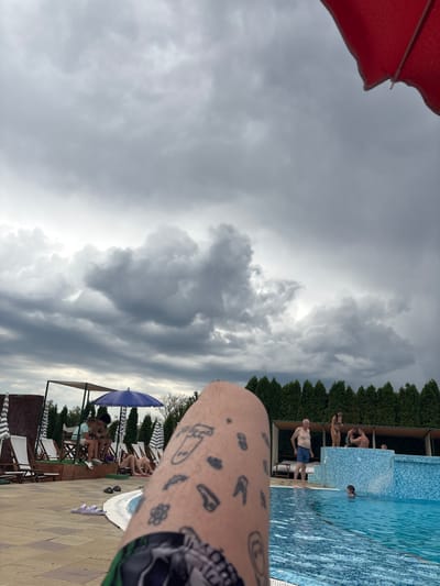The image depicts an outdoor swimming pool area under a cloudy sky. In the foreground, a person's tattooed leg and patterned shorts are visible. The pool has blue tiled walls and water. Several people are present around the pool: some are standing, some are sitting on lounge chairs or benches under shaded structures, and a few are in the water. There are umbrellas and trees in the background, with a dense line of evergreen trees forming a backdrop. The scene appears to be a leisure facility, possibly a hotel or a public pool, located in Stara Pazova Municipality, Serbia. The overall atmosphere suggests a potentially overcast or stormy day, with the clouds being a prominent feature in the sky.