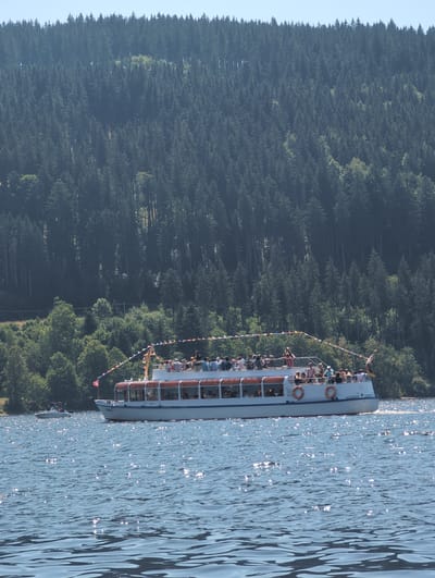 The image shows a large white passenger boat with an orange trim and many people on its upper deck, sailing on a blue, sun-dappled lake. The boat is decorated with colorful triangular flags strung along its railing. In the background, a dense forest covers a steep hillside, with a clear blue sky above. A smaller white boat is visible in the distance to the left of the passenger boat. The scene is likely located in Titisee-Neustadt, Germany, on the Titisee lake, known for its passenger boat tours amidst forested hills.