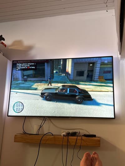 A television is mounted on a white wall, displaying gameplay from Grand Theft Auto IV. The scene shows a character standing outside a building, with a black car nearby. In-game text is visible at the bottom, providing instructions for phone calls. To the right of the TV, a wooden post with multiple electrical outlets and various plugs is visible, accompanied by a mess of black cables. The top right corner reveals a person's bare feet.