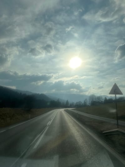 A wide-angle landscape photograph captured from a vehicle displays a two-lane asphalt road traversing a rural, mountainous environment under a partially cloudy sky. The bright sun, positioned in the upper right quadrant, casts significant glare and lens flare across the scene. The road, featuring a double white line with a dashed inner line, extends from the bottom center of the frame and curves gently left into the middle distance, appearing wet or highly reflective. To the right of the main road, a narrower paved path runs parallel. A triangular warning sign, white with a red border and a black upward-curving arrow indicating a right bend, stands on a metal post beside this parallel path on the right side of the image.
On the left side of the road, a strip of dry, light brown grass leads to a dense, dark green forest covering the lower slopes of hills and mountains that recede into the hazy background. On the right side, a grassy field slopes upwards, interspersed with sparse, bare deciduous trees and some evergreens further back, revealing more distant, cloud-shrouded mountain ranges. The sky, occupying the upper two-thirds of the image, is filled with scattered cumulus clouds, ranging from light grey to white. No individuals or other vehicles are visible. The scene is located in Sankt Georgen am Kreischberg, Austria.