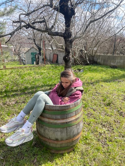 A young female is seated within a weathered wooden barrel in a grassy outdoor area in Zdravets, Bulgaria. She wears a maroon hooded sweatshirt, a khaki green puffer vest, light green leggings with a black Nike swoosh on the left calf, white Nike crew socks, and light-colored athletic shoes with multi-toned accents. Her head is angled downwards towards her crossed forearms, which rest on her lap. The barrel is constructed of wooden staves secured by rusty and green-painted metal bands. Behind the subject, a mature, deciduous tree with dark, gnarled bare branches stands. Further back, a wire fence separates the immediate foreground from several small, single-story utility structures or sheds. One structure features a dark green door, and another, positioned further to the right, has a reddish-brown door. The ground is covered in green grass with sparse patches of dry vegetation. The sky overhead is predominantly blue with scattered white cumulus clouds, under bright daylight conditions.