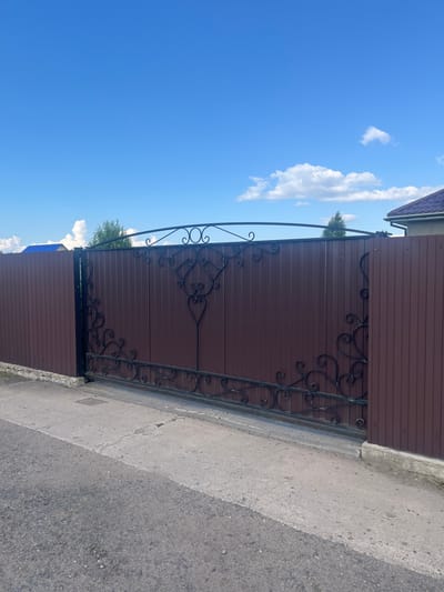 The image shows a section of a brown metal fence with decorative wrought iron elements. The fence appears to be a gate, with a black wrought iron arch extending over the top and intricate scrollwork decorating the panels. The fence is positioned next to a grey paved surface, likely a sidewalk or driveway, which then transitions to an asphalt road. In the background, a clear blue sky with a few scattered white clouds is visible, suggesting fair weather. A green tree is partially visible behind the fence. The scene is outdoors, and the lighting indicates it is daytime. The location context suggests this is a rural or suburban area in Belarus. No people or specific activities are depicted, and there is no visible text in the image. The overall impression is of a well-maintained property entrance.