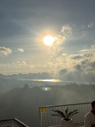 The image captures a sun-lit sky with scattered clouds. A landscape including trees, mountains, and water are in the background. A balcony with a person, a plant in a pot, and metal railings are in the foreground.

While the location is not explicitly determined to be Kedisan, Indonesia, it cannot be confirmed or denied.