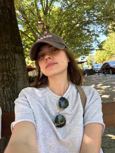 A young woman with long brown hair and blue eyes, wearing a gray sweatshirt, a brown baseball cap with pink lettering, and sunglasses perched on her shirt, is seated outdoors. The background reveals trees with dappled sunlight and a glimpse of cars suggesting a street scene. The location is inferred to be Siófok, Hungary.
