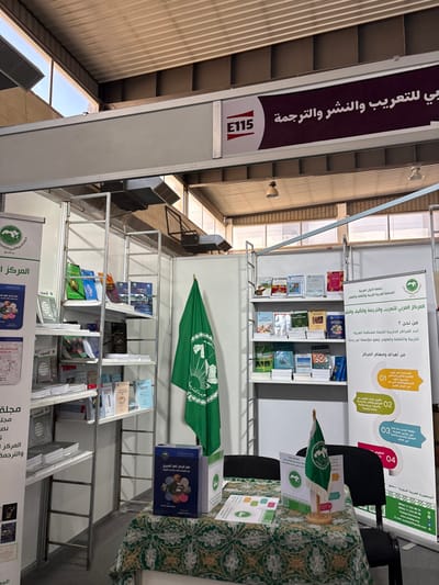 The image displays an unoccupied exhibition booth, identified by the overhead sign "E115," located indoors within the city of Al-Ghizlaniya, Syria. The booth is dedicated to "The Arab Center for Arabization, Publishing, and Translation," as stated on a prominent horizontal sign and detailed vertical banners. Two tall, multi-shelf metal units are fully stocked with numerous books of varying sizes and cover designs. A large green flag with the center's circular logo and Arabic text hangs vertically within the booth. A rectangular table in the foreground is draped with a green patterned tablecloth and features a large blue-covered book, a small desk flag with the same logo on a wooden stand, and several white brochures. Two black chairs are partially visible behind the table. The surrounding environment includes a high, corrugated metal ceiling with visible skylights and metal ventilation ducts. No individuals are present in the scene.