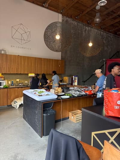 This image shows the interior of a business, likely a restaurant, in Culver City, California. Two large, spherical pendant lights hang from the ceiling. A counter with multiple people preparing food stretches across the length of the image. The counter is lined with trays containing various ingredients. A dog is lying on the floor near the end of the counter. Customers are standing by the end of the counter, with one holding a red shopping bag. A sign with "EM COSMETICS" is visible on a white wall next to a wooden cabinet.