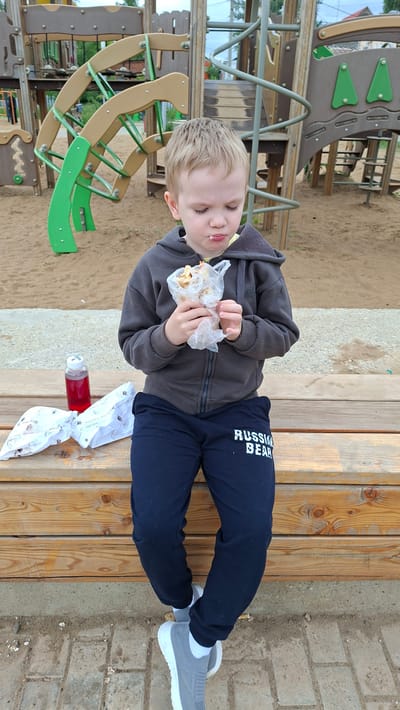 A young boy with blonde hair is sitting on a wooden bench in a playground in Nytva, Russia, eating a food item wrapped in plastic. He is wearing a dark gray hooded sweatshirt and dark blue sweatpants with "RUSSIA BEAR" printed on the left leg. He has white socks and gray sneakers. To his left on the bench are two white wrappers, one of which appears to be for a food item, and a small red bottle. The playground equipment, consisting of a colorful, multi-level structure, is visible in the background. The ground is covered in sand and brick pavers.