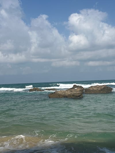 This is a photograph of the Mediterranean Sea near Shoshanat HaAmakim, Israel. The image shows a rocky coastline with several large rocks protruding from the turquoise water. Waves are crashing against the rocks and the shore, creating white foam. The sky is partly cloudy with patches of blue visible. There are no people or animals in the frame. The scene depicts a natural coastal environment.
