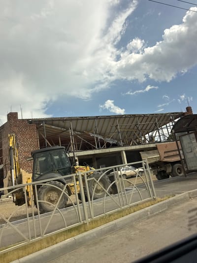 This media file captures a construction site under a cloudy sky, likely during the daytime. The scene is dominated by the skeletal framework of a building under construction, with visible metal beams and a partially covered roof. Brickwork forms a lower section of the structure, suggesting it's in an early to mid-stage of development.
A yellow excavator is positioned prominently on the site, with its cab visible. Large tires from what appear to be heavy-duty vehicles, possibly the excavator itself or other construction machinery, are also a notable detail. A white car is parked nearby, partially obscured by the construction. A metal fence or barrier runs along the side of the scene, separating the construction area from a paved surface, likely a road or sidewalk.
The sky is filled with large, fluffy clouds against a bright blue background, indicating fair weather. The overall impression is one of ongoing work and development in an urban or semi-urban setting. No people are clearly visible in the frame. There is no visible text in the image. The context provided suggests the location is Rozhdestvenskaya, Russia.