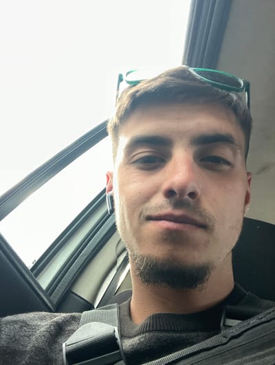 A close-up selfie of a young man with a light complexion, brown hair, and a trimmed beard. He is wearing a gray sweater and a seatbelt. Green sunglasses are perched on top of his head. The background shows the interior of a car with a window. The location is likely Yoakim Gruevo, Bulgaria.