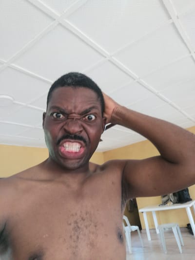 A shirtless Black man with dark hair and a mustache makes a fierce facial expression, baring his teeth and furrowing his brow. He is indoors, with a white tiled ceiling visible above. His right arm is raised, with his hand resting on the back of his head. The background includes a yellow wall, a white table, and white chairs. The scene appears to be in a residential setting in Ajasse Ipo, Nigeria. The time of day is not explicitly indicated, but the indoor lighting suggests it is daytime. The man's expression conveys anger or intense emotion. There is no visible text in the image.