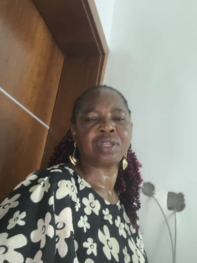 A woman is visible in an indoor setting in Mopo-Akinlade, Nigeria. She is wearing a black dress with white floral patterns and gold earrings. Her hair is styled in dark red, curly braids. The background includes a white wall with electrical conduit visible, and a wooden door frame.
