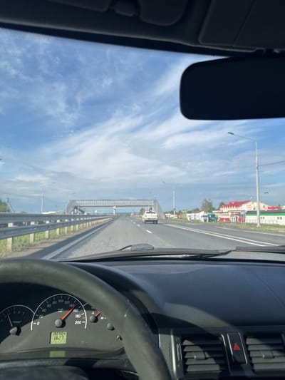 The image captures a driver's perspective from inside a car traveling on a road in Усть-Сыны, Russia. The view through the windshield shows a multi-lane asphalt road stretching into the distance, bordered by a metal guardrail on the right. Beyond the guardrail, what appears to be a body of water or a wide expanse with a distant bridge structure is visible, leading to buildings with colorful rooftops (red and green) and some industrial-looking structures further along the roadside. Overhead, the sky is bright blue with scattered, wispy white clouds. Several streetlights/power poles line the road.

Inside the car, parts of the dashboard and steering wheel are prominent. The black steering wheel occupies the lower right portion. To its left, the instrument cluster is visible, showing a speedometer indicating approximately 80 km/h. An odometer displays "190904" km, and a digital clock shows "18:27". Other text on the speedometer includes "km/h" and "VDO". Above the dashboard on the right, air vents and a red triangular hazard light button are visible. A rearview mirror and part of a sun visor are visible in the upper left, reflecting the sky. The time of 18:27 combined with the bright sky suggests late afternoon or early evening on a clear, sunny day. The scene depicts typical travel on a rural or intercity road, possibly crossing a bridge over water, at a moderate speed.
