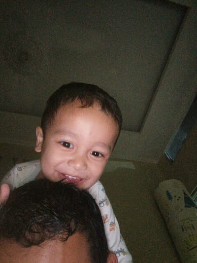 In a dimly lit indoor setting, a young child with dark hair and brown eyes smiles at the camera, their mouth open and tongue visible. The child is wearing a white onesie with a cartoon pattern. The child's head is resting on the head of an adult, whose dark hair is visible in the foreground. The background features a textured ceiling and a corner of a room. The image appears to be taken in Cibungbulang, Indonesia, likely in a residential area given the casual indoor environment.