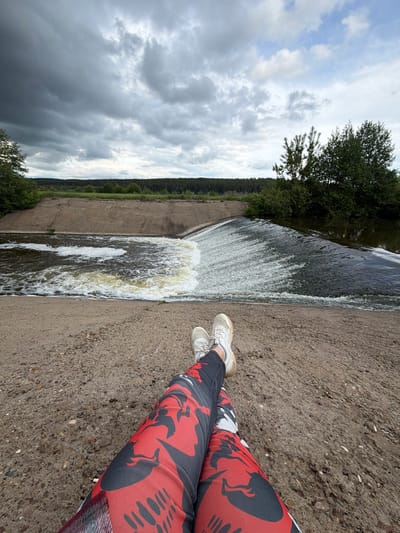The photograph presents a scene at the location of the dam in Nizhnii Vishur, Russia. The view is from a high angle, looking down at the dam and the water flowing over it. An individual is present, with their legs extending into the frame, clad in red and black patterned pants and wearing white sneakers. The dam is constructed of concrete, and water is rushing over the edge. Beyond the dam, a line of trees and dense vegetation is visible along the riverbank. The sky is overcast with dark clouds.