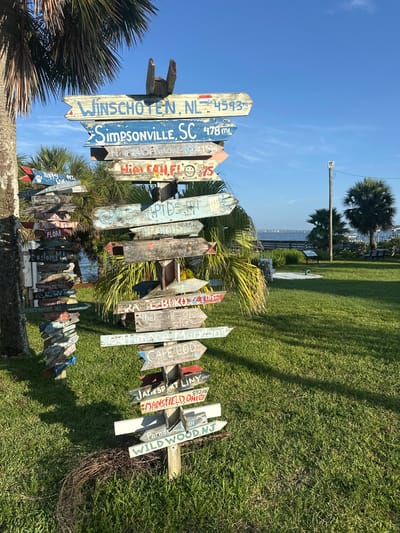 The image shows a signpost with multiple directional signs pointing in various directions, indicating different cities and distances. The signpost is located in an outdoor grassy area, with palm trees and a body of water in the background. The sky is clear and blue. The signs are weathered and appear to be made of wood, with hand-painted text. Some of the visible destinations include Winschoten, NL; Simpsonville, SC; and High Falls, FL. The signpost is located at a park in Melbourne Beach, Florida, United States. There are no people or animals visible in the image. The overall environment is sunny and appears to be a recreational area.