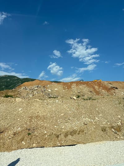 The image depicts a construction site under a clear blue sky with scattered white clouds. The scene is dominated by a dirt embankment, heavily laden with rocks and gravel, suggesting ongoing excavation or landscaping work. A bright yellow excavator is visible on the embankment, partially obscured by the terrain, hinting at active construction. A smaller green vehicle is also present on the site, positioned lower down the slope. In the background, a lush green, tree-covered mountainside rises, providing a stark contrast to the barren construction area. The sky is a vibrant blue, indicative of a sunny day, likely mid-morning or afternoon given the lighting. The lower right portion of the image shows a section of white gravel, possibly a pathway or prepared area for construction materials. The overall impression is one of an active, outdoor construction project in a hilly or mountainous region. There is no visible text in the image.