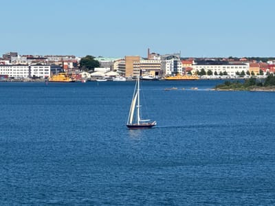 A wide shot captures a clear, blue sky above a cityscape on the coast of Karlskrona, Sweden. The city features a collection of buildings with varied architecture, including residential blocks, commercial structures, and industrial elements. A prominent yellow ferry is docked near a building with large windows. In the foreground, a sailboat with white sails navigates the blue water. The scene is set on a sunny day, with calm water reflecting the sky.