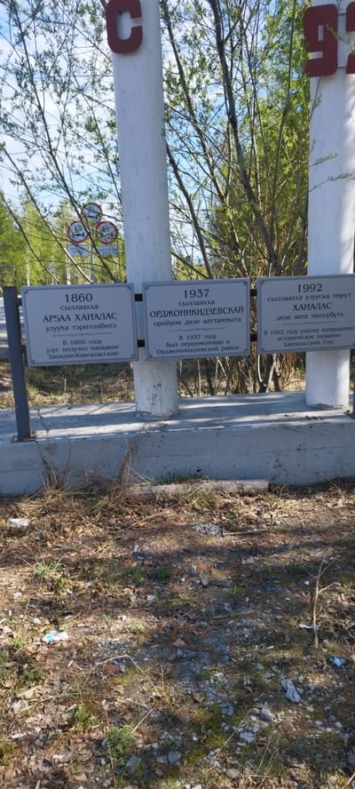 The image shows three commemorative plaques displayed on pillars, likely marking historical events. The plaques are silver in color and have decorative borders. Each plaque contains information in both the local language and Russian. The first plaque is labeled "1860" and refers to the formation of the Arbaa Khanalas ulus (district) and the renaming of the district to Zapadno-Kangalassky. The second plaque is labeled "1937" and mentions the renaming of the area to Ordzhonikidzevsky district. The third plaque is labeled "1992" and notes the return of the historical name Khanalas. The pillars holding the plaques are concrete and painted white. The scene is located in Tabaga, Russia, as indicated by the Russian language on the plaques. The background contains greenery, including trees and shrubs. The foreground consists of dry grass and ground debris. The presence of these plaques suggests the location is a place of historical significance within the community.