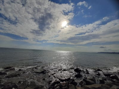 The image depicts a rocky shoreline in Atakum, Turkey, with the Black Sea extending to the horizon. The sky is partly cloudy, with the sun partially obscured by clouds, casting a bright reflection on the water. Waves are gently lapping against the shore, where dark, irregular-shaped rocks are visible. In the distance, a city skyline is faintly discernible along the coast. There are no people or animals present in the image. The scene conveys a sense of natural beauty and tranquility.