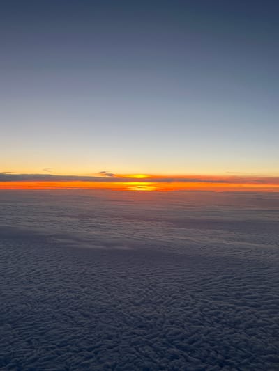 The image depicts a sunrise or sunset observed from above the clouds. The sky transitions from a dark blue on the left to a lighter hue near the center, where the sun is partially obscured by a layer of clouds. This cloud layer is illuminated with orange and yellow hues from the sun. Below this illuminated layer is a dense expanse of dark, textured clouds that fill much of the right side of the image. There are no people or identifiable objects. The location is not discernible from the image content.