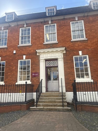 The image shows the exterior of a brick building in Thrapston, United Kingdom. The building has multiple white-framed windows on the facade, and a prominent white door frame around the entrance. There are stairs leading up to the door, flanked by black iron railings. A purple sign reading "Bletsoes Est. 1881" is visible next to the entrance. The building is likely a commercial or institutional structure. The adjacent sidewalk is paved with bricks.