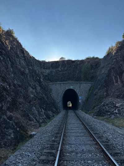 This is a low-angle, wide shot of train tracks leading into a stone train tunnel. The tunnel entrance is arched and constructed from rough-hewn stones, with a smaller, arched opening visible within the larger archway. Light streams from the tunnel's exit, silhouetting the interior. The tracks are made of steel rails and wooden ties, laid on a bed of gravel. On either side of the tracks, steep, rocky hillsides rise sharply. The left hillside is covered in dark, rough rock with sparse vegetation. The right hillside also consists of rock and vegetation, with some areas reinforced with netting. The sky above is clear and a pale blue, with the sun low on the horizon, casting a warm glow. The overall environment suggests a remote, natural setting. This location is the Tunnel de Générargues, located in Générargues, France.