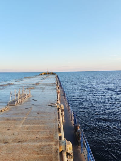 The image shows a view from a boat or pier on a calm body of water. The foreground features the weathered, rusty surface of the deck, with visible circular hatches and a white metal walkway with railings. A blue railing lines the edge of the deck, separating it from the dark blue water. In the distance, the deck extends towards a point with yellow bollards and a structure resembling a small tower. The horizon is clear, with a pale blue sky above. The scene appears to be located in Kakkarovo, Russia, given the context. There are no people visible in the image, and the overall impression is one of stillness and expanse.