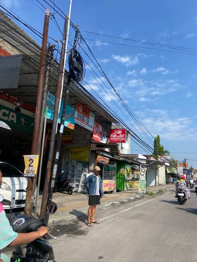 The image shows a street scene in Kecamatan Tulangan, Indonesia (latitude: -7.481, longitude: 112.646). The street is lined with shops and businesses. There are many power lines overhead. A man in a blue denim jacket and black shorts is standing on the sidewalk, facing the street. He is looking to his left. He is standing in front of a shop with a sign that reads "GORENGAN".  A motorcycle is parked on the sidewalk to his left, and a car is parked to his right. There is a motorcycle in the street, which is empty. The sky is bright blue with a few clouds.