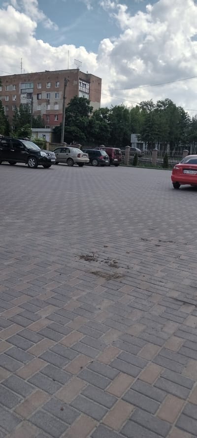 The image depicts a paved parking lot in front of a multi-story residential building. The building, which appears to be constructed of brick and concrete, has multiple windows and balconies, with a menorah visible on its roof. Several cars are parked in the lot, including a dark SUV, a light-colored sedan, a dark hatchback, and a red sedan. Trees and a fence are visible behind the parking lot, separating it from other structures. The sky is cloudy. The specific location is the city of Valky, Ukraine.