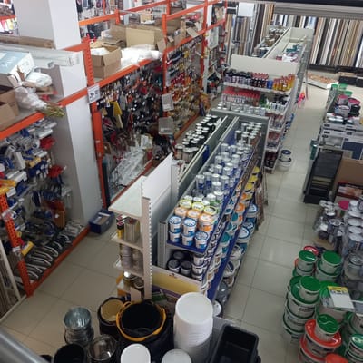 The image shows the interior of a store, likely a hardware or home improvement store, in Briceni, Moldova. The store is filled with shelves stacked with various products, primarily paint cans, buckets, and painting tools like rollers and brushes. The products are neatly organized, with many paint cans in different colors and sizes displayed on blue shelves. Some boxes and packaging are also visible, along with some tools like pliers and hammers. The lighting suggests it is daytime, and the overall atmosphere appears to be that of a retail environment. Visible text includes brand names like "KNAUF," "POLIGHT," "MTK," "MAFLEX," and "GRUNT," as well as "SUPER FINISH" and "PRIMAGOL." There are no people visible in the image. The store appears to be well-stocked and organized, catering to customers looking for painting and DIY supplies.