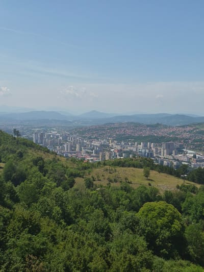 This is an aerial, eye-level photograph taken from the MZ Trebević, Bosnia and Herzegovina, looking out over a city and surrounding hills under a clear, pale blue sky. In the foreground, a lush green, tree-covered hillside slopes downwards. The mid-ground is dominated by a dense urban landscape featuring numerous tall, modern buildings, including many apartment blocks and some commercial structures. Further in the distance, rolling hills covered in residential housing and vegetation stretch towards the horizon, with hazy, layered mountains forming the backdrop. The overall impression is of a city nestled within a verdant, hilly environment.