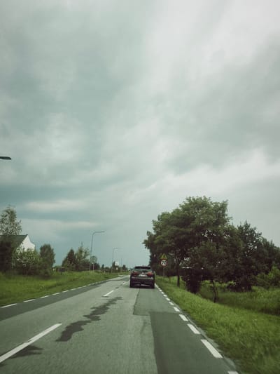 An overcast sky looms above a two-lane asphalt road in Krubin, Poland. The road curves slightly to the left, with wet patches visible on its surface. Green grass lines both sides of the road, with trees and some buildings visible in the distance. A dark SUV is driving ahead on the right lane, with a speed limit sign showing "70" to its left. The image appears to be taken from a moving vehicle, capturing a moment on a rural road under a cloudy sky.