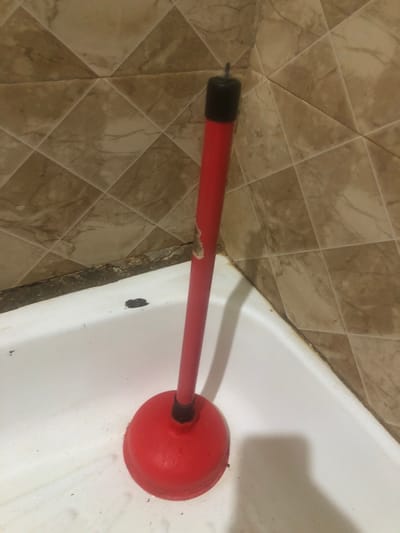 A red toilet plunger with a black cap rests inside a white bathtub. The plunger's handle is red, and its rubber cup is also red. The plunger is positioned vertically in the corner of the bathtub, with the cup resting on the bottom and the handle extending upwards. The bathtub appears to be made of porcelain or a similar material and is in a state of disrepair, with visible grime and a damaged seal along the edge. The wall behind the bathtub is tiled with beige, diamond-shaped tiles. The overall scene suggests a domestic bathroom setting.