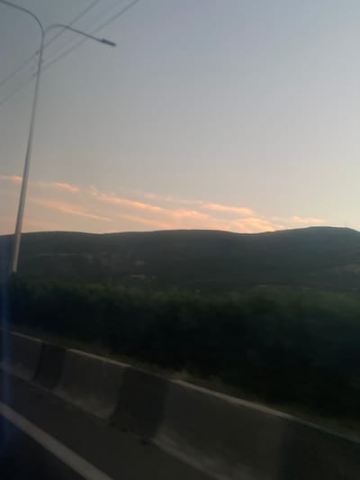 A wide shot captures a dusky sky over a dark, rolling hillside, with a concrete barrier and a road in the foreground. The sky is a pale gradient of blue to light gray, with faint, elongated pink clouds near the horizon. A tall, thin lamppost with wires is silhouetted against the sky on the left side of the frame. The hillside is covered in dark vegetation, appearing almost black in the dim light. In the immediate foreground, a concrete barrier with angled sections runs diagonally across the bottom of the image, with a white line marking the edge of a road to its left. The scene appears to be from a moving vehicle, likely on a highway.