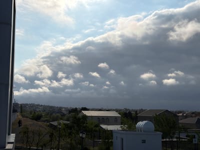 The image presents a view of Sumqayit, Azerbaijan, under a partly cloudy sky. The foreground contains foliage and architectural elements including a white, rectangular building with a spherical white object on top, likely a water tank. The middle ground reveals a cluster of buildings and residences typical of a suburban or urban-rural setting. A larger, light-colored building with multiple arched windows is visible amidst the trees. Distant views show the cityscape stretching towards the horizon. A building or a construction is seen on the left side of the image.