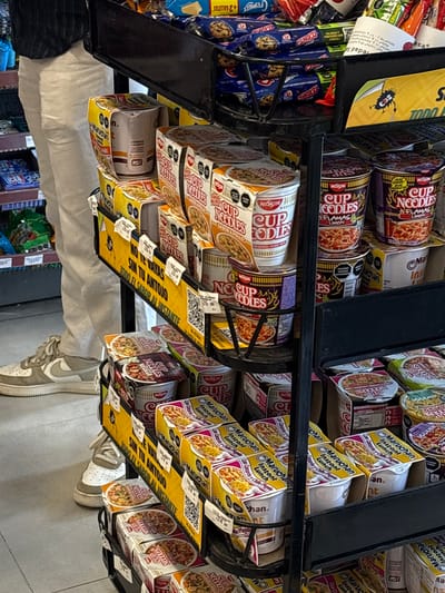 The image depicts a close-up view of a shelf stocked with instant noodle cups, predominantly Nissin Cup Noodles and Maruchan Instant Lunch, in a retail environment. The products are arranged in multiple tiers on metal shelving units. In the background, a person is partially visible, wearing light-colored pants and white sneakers, suggesting they are a shopper. The overall setting appears to be a convenience store or a section of a larger market. The lighting is adequate, illuminating the products clearly. The specific location of San Juan Tejaluca, Mexico, is not discernible from the visual information present in the image itself.