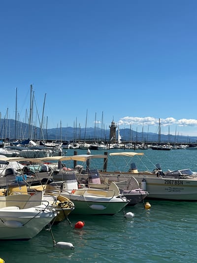 A view of the harbor in Desenzano del Garda, Italy, showcases a bright, clear day with a vast blue sky. The foreground is dominated by several boats docked side-by-side. These include a white motorboat on the left, a smaller green boat next to it, and a larger white boat with the registration "2067 BSN" and "BERTOLDI BOATS" visible on its side. Various buoys, both white and red, are floating in the turquoise water, tethered to the boats.

In the mid-ground, numerous sailboats with tall masts fill the harbor, creating a dense forest of rigging. A stone lighthouse with a lantern room stands prominently among the sailboats, positioned near the far end of the docked vessels. The background features a range of distant mountains, some with a hazy appearance, under the bright sun. A breakwater wall, lined with more sailboats, separates the harbor from the open water. The overall scene is one of a busy, sunlit marina on a calm day.