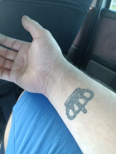 The media file shows a close-up view of a person's left forearm and hand, with a black tattoo on the wrist. The tattoo appears to be a stylized representation of brass knuckles with a crown design. The skin is fair and shows some light hair growth. The person is wearing a blue t-shirt, visible in the lower part of the frame. The background is out of focus but suggests an indoor setting, possibly a car interior, with dark upholstery and a glimpse of what might be a seatbelt. The lighting appears to be natural, suggesting daytime. There is no visible text in the image. The image is cropped to focus on the forearm and tattoo.