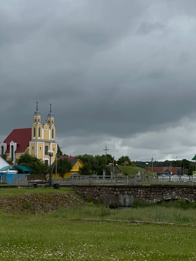 The image depicts a village scene in Belarus under a heavy, overcast sky. The main subject is a striking yellow church with two tall, grey, domed towers topped with crosses. It has a red roof and white-trimmed windows. Adjacent to the church, a smaller yellow building with a single window is visible.

The scene is viewed from a slightly elevated grassy area, which slopes downwards towards the church. A railway track with a stone embankment runs horizontally across the middle ground, separating the church from the foreground. A metal fence runs along the edge of the railway. In the foreground, a lush green lawn is dotted with small white flowers.

Further in the background, more buildings with red roofs and green trees can be seen, suggesting a small rural settlement. A single street lamp is visible near the church. The overall weather appears to be stormy or very cloudy, casting a somber mood over the picturesque village. The time of day is indeterminate due to the cloudy conditions. There is no visible text in the image.