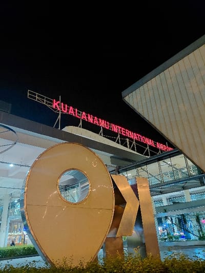 The image depicts the exterior of Kualanamu International Airport, situated in the city of Sidourip, Indonesia, at night. In the foreground, a large, metallic, three-dimensional sculpture resembling a geographical pin icon, with a circular cutout, stands amidst low green foliage. To its right, partially visible, are blocky metallic letters appearing to form "KNO." Behind this sculpture, the modern airport building is illuminated, featuring large glass facades and an elevated glass-enclosed walkway. Mounted high on the building's structure, a prominent red neon sign displays the text "KUALANAMU INTERNATIONAL AIRPORT." The sky above is dark, indicating nighttime, with artificial lighting from the airport structure and the neon sign providing illumination to the scene. No discernible individuals or specific activities are visible in the immediate foreground, though distant interior lights suggest activity within the airport building.