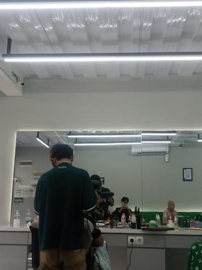 The image depicts the interior of a barbershop or salon located in Sindanglaya, Indonesia. The scene is brightly lit by multiple horizontal LED strip lights affixed to a white corrugated metal ceiling. A security camera is mounted on the upper left wall.
The primary focus is a male individual, seen from behind, standing in the foreground, wearing a dark green short-sleeved polo shirt with "PIXEL" visible on the back collar, and light brown trousers. He appears to be a barber, attending to a client whose head and shoulders are partially visible beneath a dark cape. The client is seated in a barber chair directly in front of the barber.
A large mirror covers the central and right portions of the wall, reflecting the room. In the mirror's reflection, the barber is visible facing the client, holding what appears to be hairdressing equipment. To the right of the reflected barber, a second male is visible, holding a dark object, possibly a phone or camera, at chest height. Further to the right in the reflection, a female individual wearing a light-colored hijab and top is seated, observing the scene.
The counter below the mirror holds various bottles, possibly hair products, and hairdressing tools such as brushes and clippers. Electrical outlets are visible on the counter. An air conditioning unit is reflected on the wall above the mirror, and a framed document or sign is also reflected on the right wall. A Wi-Fi symbol sticker is visible on the mirror. The walls are painted a light, neutral color.