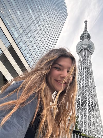 A low-angle, full-body selfie shows a young woman in the foreground smiling at the camera, her face partially obscured by her long, wavy, light brown hair. She is wearing a dark blue jacket. Behind her, to the left, is a modern glass-fronted skyscraper, and to the right, the Tokyo Skytree, a tall, lattice-like observation and broadcasting tower. The sky is overcast. The image appears to have been taken outdoors in the city of Sumida, Japan, given the presence of the Tokyo Skytree.