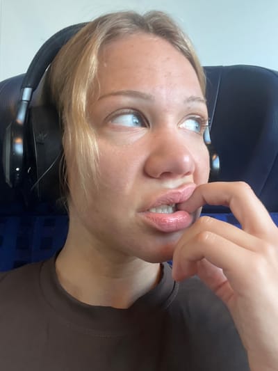 A close-up, waist-up shot shows a young woman with blonde hair wearing black headphones, a dark brown t-shirt, and a light pink lipstick. Her head is turned to her right, and she is looking off-camera, with her right thumb and index finger biting her lower lip. The background consists of a dark blue headrest of a seat, possibly in a vehicle or airplane. The overall lighting is bright and appears to be natural. The image was captured in Ampfing, Germany.