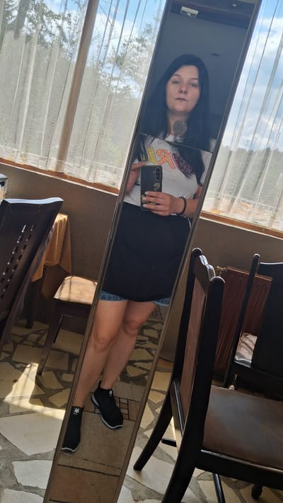 The media file is a mirror selfie taken by a young woman with long, dark hair. She is wearing a light-colored t-shirt with a graphic print, a black skirt, and black sneakers. In her right hand, she is holding a smartphone, which she is using to take the photo.

The woman is standing indoors, likely in a room with large windows covered by sheer curtains, allowing natural light to filter through. Outside the windows, trees and a cloudy sky are visible, suggesting it is daytime. The floor appears to be tiled. A wooden chair with a dark finish is visible in the background, reflected in the mirror. The overall setting suggests a relaxed, possibly domestic environment.

The woman's expression is neutral, and she appears to be looking directly at her reflection. There are no visible signs of any specific event or activity, beyond the act of taking a selfie.

There is no discernible text in the image.

The photo was taken in Marchevo, Bulgaria.
