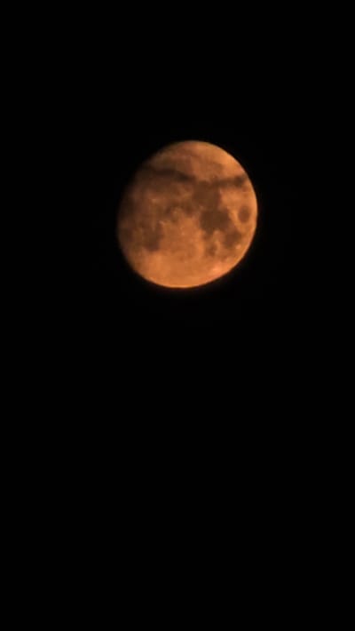 A close-up, vertical shot shows a full moon against a black sky. The moon is a deep orange color and its surface is textured with darker patches that suggest craters and maria. The illumination of the moon is uneven, with one side appearing brighter than the other, indicating the direction of the light source. The overall impression is of a celestial object observed at night.